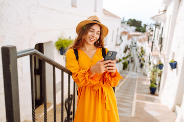 Young tourist in a hat and a bright dress with a phone sits on the steps in a picturesque town. A...