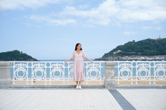 Chica joven morena con vestido rosa de verano apoyada sobre la barandilla del paseo mar&iacute;timo en la playa de la Concha, San Sebasti&aacute;n (Donostia) - Espa&ntilde;a: sin mirar a c&aacute;mara