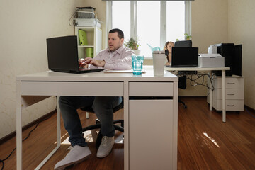 Productive home office moment featuring a focused man juggling a phone call and computer work, while his female teammate reaches for the printer tray beside her organized desk setup