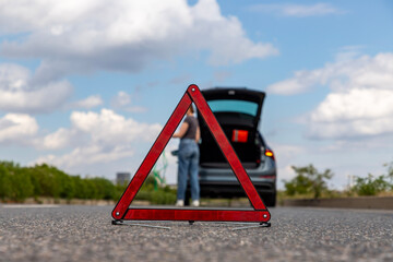 Roadside emergency: woman waiting for help with warning triangle in front of broken car under bright blue sky