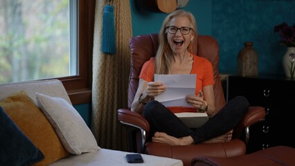 Portrait of mature woman in a red top and glasses sits in a brown leather recliner, reading a letter and smiling broadly. Her feet are bare and she is looking at the letter with a joyful expression.