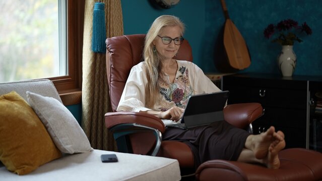 Using glasses to see tablet computer, a mature woman with blonde hair sits in a brown leather recliner. Her feet are propped up on the footrest and she has a relaxed smile. 
