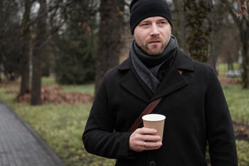 Pensive hipster drinking coffee in the urban street. Handsome man wearing black coat, brown leather...