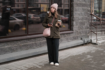 Portrait of mature woman in wool hat standing on the urban street, drinking takeaway coffee from paper cup. Middle-aged 40s female walking around the city at cold autumn day. Living in modern district