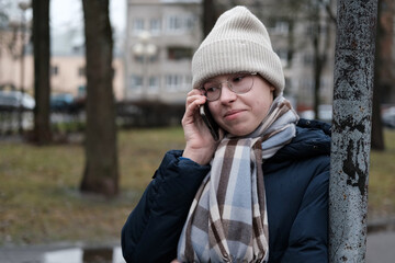 Teenager Girl Walking Alone on City Street with Smartphone. Portrait of 14 years old girl wearing eyeglasses, wool hat, winter jacket using mobile phone. Urban Style. Rainy mood weather. Cold season