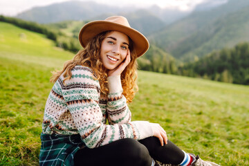 Young female traveler enjoying mountain scenery standing on green hilly meadow. Beautiful woman in hat feeling freedom outdoors. Travel, nature, hiking concept.