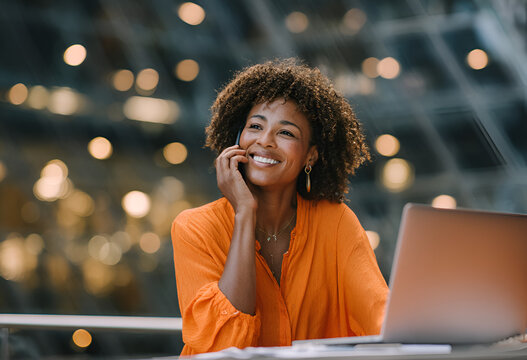 Smiling woman with curly hair talks on her phone while sitting in front of a laptop