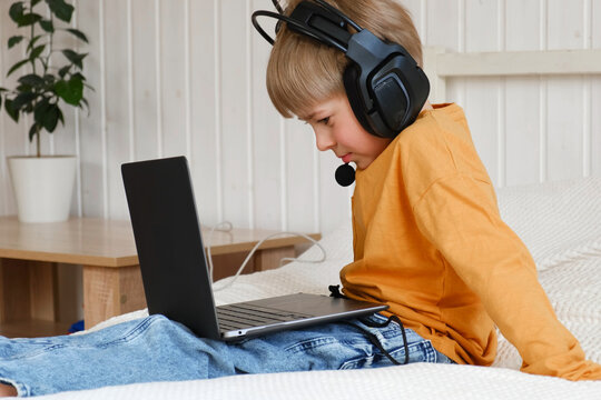 Little smiling 6 years old boy using a laptop with headphones. Child having video call with friends, granny at home in bed. Kid looking at display and waving hand. Childhood leisure activity concept