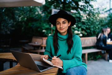 Brunette freelancer sits at table in leafy outdoor cafe, casually dressed, writing in planner next to laptop, conveying balanced approach to remote work and personal reflection.