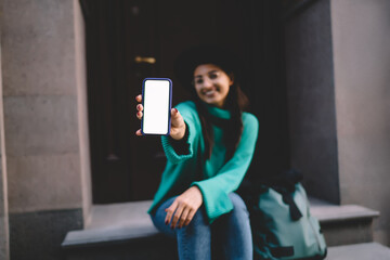 Playful woman showing smartphone screen toward camera while sitting on stairs, blank white display...