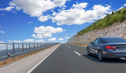 Modern black coupe driving on a scenic highway beside a rocky hillside under a bright blue sky with...