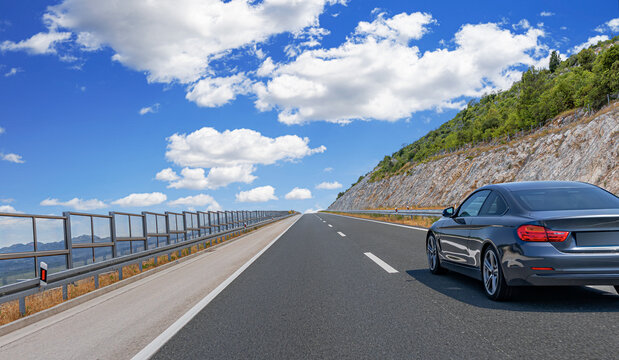 Modern black coupe driving on a scenic highway beside a rocky hillside under a bright blue sky with fluffy clouds.