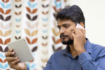 Angry or Sad Asian Indian man using multiple digital devices, smart phone and tablet to make a call and browsing to multi task