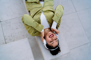 Young woman lying on modern bench, enjoying music through wireless headphones. Her joyful...