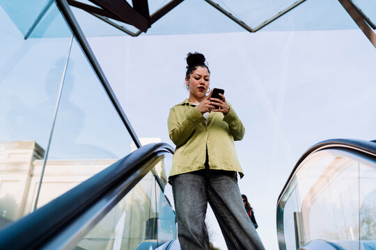 Young Hispanic woman on escalator purchases tickets via a mobile app. Focused on her screen, Female efficiently books travel or events, using technology to streamline her daily plans on the go
