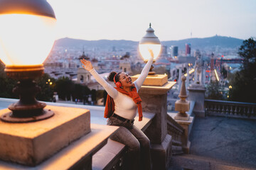 Cheerful young woman with wireless headphones enjoys music at sunset, sitting on a historic balcony with arms raised. She embraces city skyline, feeling free, joyful, and inspired by the evening view