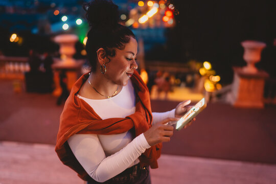 smiling woman enjoys digital experience on her smartphone at night. City lights reflect in her eyes as she browses online, highlighting modern connectivity, social media, and mobile engagement