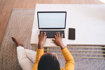 Cheerful African woman in glasses working on laptop, sitting on floor by coffee table in cozy home interior, smiling with joy while engaging in remote freelance work