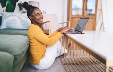 Happy African woman in glasses working on laptop while seated by coffee table, relaxed and smiling...
