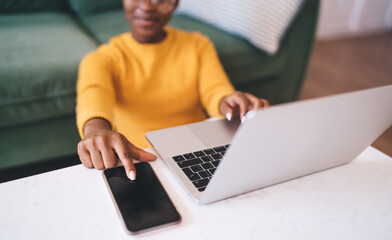 African female freelancer multitasking with laptop and smartphone at coffee table, reaching for...