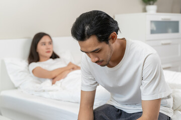 Obraz premium Young Asian unhappy girl sitting on the edge of the bed, against her boyfriend, lying in bed.