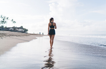 Fit young woman strolls silently along ocean edge, facing away, in dark sportswear with ponytail...