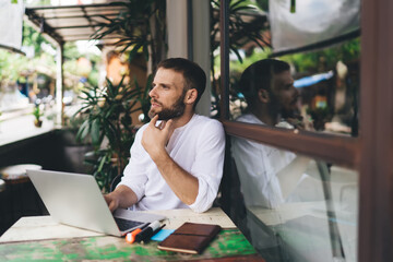 Bearded freelancer pausing in reflection, touching chin, looking away from screen. Scene...