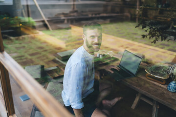 Freelancer seen through window working on laptop, symbolizing freedom of location-independent tech...