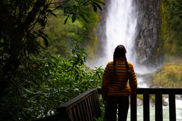 Girl stands on a wooden platform and admires the tall, impressive Thunder Creek Falls in Mount...
