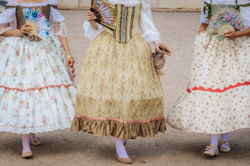 Three women dressed in ornate vintage gowns with lace and floral patterns hold decorative fans...