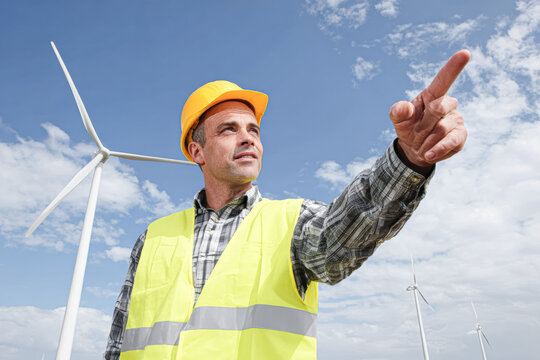 A construction worker wearing a yellow helmet and reflective vest points forward with wind turbines in the background under a partly cloudy sky.