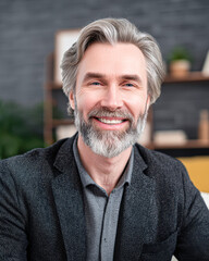 Fototapeta premium A middle-aged man with gray hair and a beard smiles warmly, dressed in a blazer and shirt, sitting in a modern indoor setting with blurred background elements.