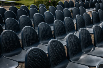 Obraz premium Multiple rows of identical black chairs are neatly arranged in an outdoor setting on a dirt surface, creating a uniform and symmetrical pattern.