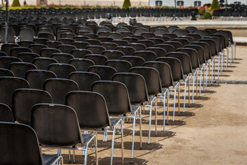Fototapeta premium Multiple rows of identical black chairs are neatly arranged in an outdoor setting on a dirt surface, creating a uniform and symmetrical pattern.