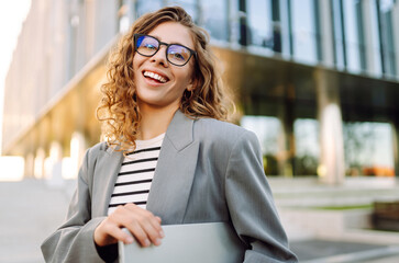 Stylish woman in glasses and with laptop posing near modern building at sunset. Beautiful female freelancer enjoying weather after work outdoors. Business and technology concept.