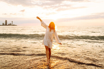 Cheerful woman in white dress enjoys sunset while walking along the seashore. Young woman feels...