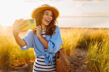 Selfie time! Happy woman on the beach with a phone enjoying the sunset on the beach. Young tourist...