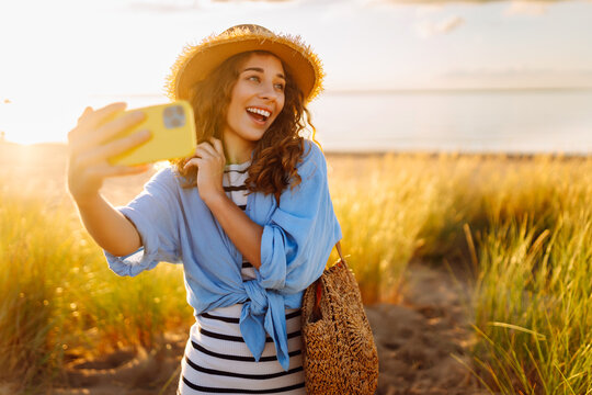 Selfie time! Happy woman on the beach with a phone enjoying the sunset on the beach. Young tourist in a hat and with a bag enjoying the moment in nature. Blogging and technology concept. - Powered by Adobe