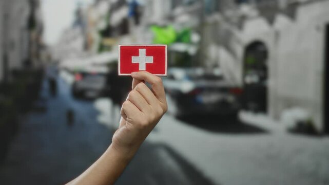 Hand of caucasian man holding switzerland flag patch on a busy outdoor street in a town, signifying swiss culture and national pride through embroidered detail.