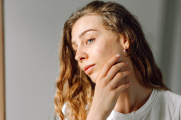 Close-up face of a young woman with rashes looking at the mirror in a cozy room. Beautiful woman...