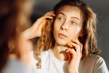 Close-up face of a young woman with rashes looking at the mirror in a cozy room. Beautiful woman...