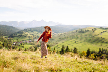 Young female traveler in a hat walking on a green hill and looking at the mountain scenery. Beautiful woman enjoying sunny weather and nature outdoors. Adventure, freedom and nature concept.