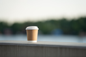A brown coffee cup is placed on a concrete ledge overlooking a calm body of water. The soft morning...