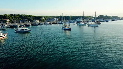 Serene harbor scene featuring multiple sailboats and yachts moored in calm blue waters with charming coastal town buildings in background. Perfect for travel content