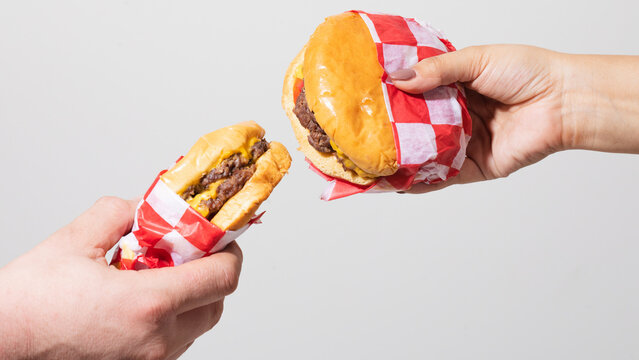 Two hands holding American cheeseburgers wrapped in red checkered paper on white background