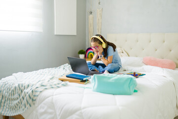 Cute latin girl doing her homework on laptop while sitting on bed