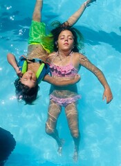 Two siblings float on their backs in a swimming pool, heads gently touching and bodies extended in opposite directions, eyes closed and facing upward.