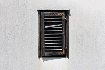 A rustic, weathered wooden window shutter with broken horizontal slats is mounted on a plain white stucco wall, evoking a sense of abandonment and vintage architectural charm
