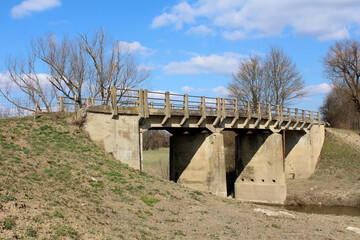 A weathered concrete bridge with sturdy pillars crosses a mostly dry riverbed, surrounded by...