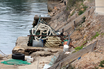 A pile of thick ropes, fishing nets, and plastic containers sits atop a cracked concrete pier,...